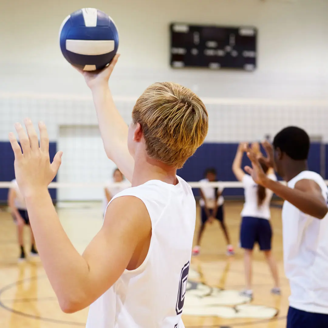 A view from behind a volleyball player in a white jersey as they toss a blue and white ball into the air for a serve on an indoor court.