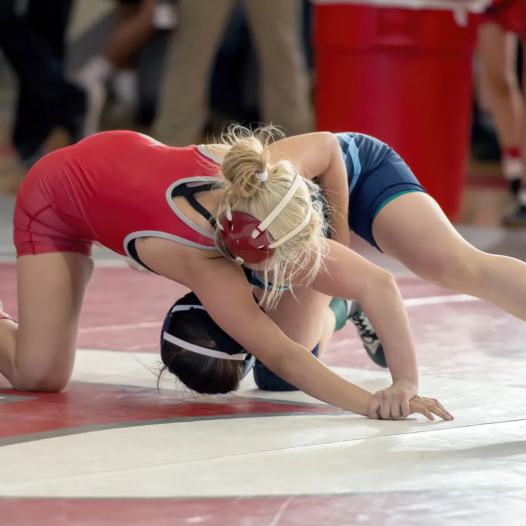Two youth wrestlers in red and blue singlets grappling on a competition mat during a match.