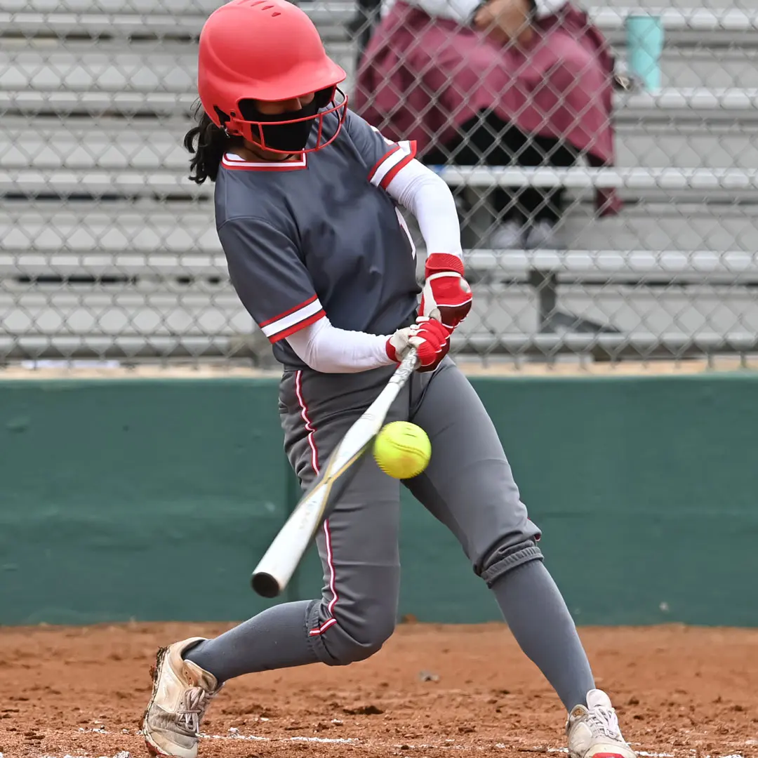 An action shot of a softball player in a grey and red uniform swinging a bat and making contact with a yellow softball on a dirt infield.