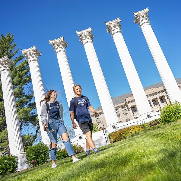 Two happy students walk uphill past five white columns on a sunny campus day.